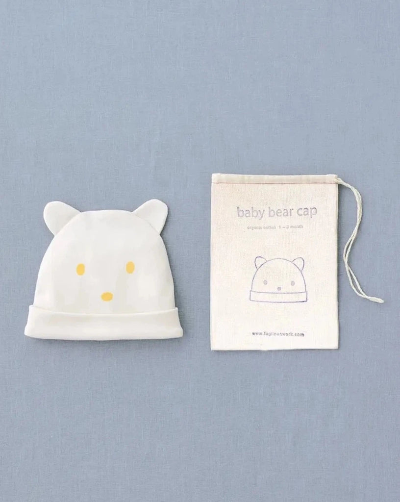 A white Baby Bear Cap with bear ears and a simple bear face design is placed next to a beige drawstring bag. The bag has "Baby Bear Cap" and a bear face illustration printed on it, making it the perfect newborn gift. The items are displayed on a light blue fabric background.