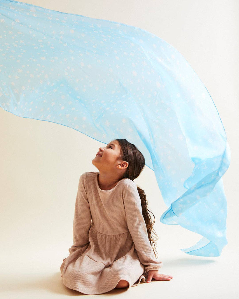 A young girl in a beige dress sits on the floor, smiling upward. Above her, Sarah's Silk Giant Snow Playsilk, a large and translucent blue fabric adorned with white floral patterns, sways gently. It invites open-ended play and ignites her creative potential in the light, airy atmosphere.