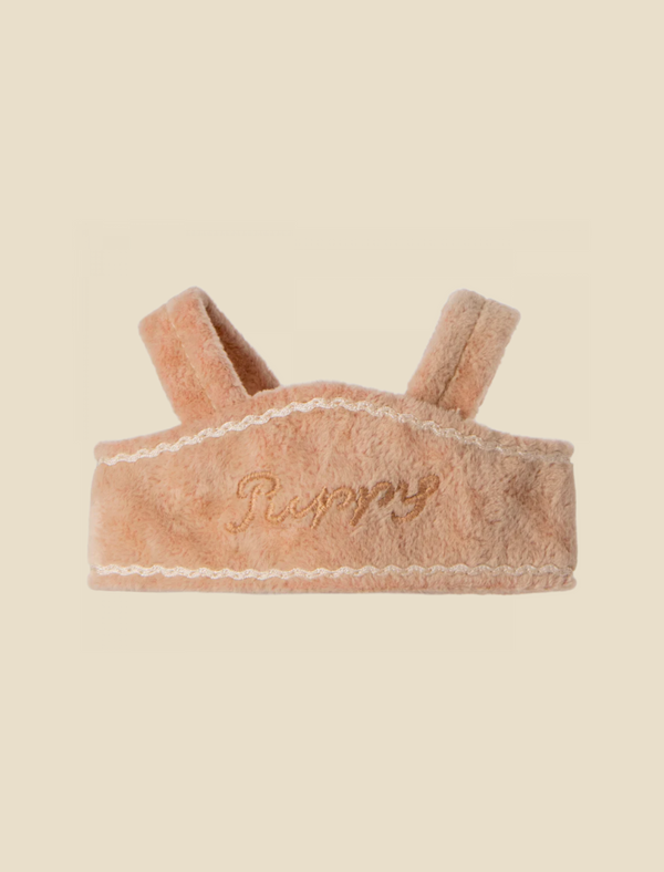 A soft, beige, textured headband with the words "Maileg Puppy Supply, Harness" embroidered in cursive on the front. It has two strap-like extensions at the top, possibly for holding it in place like a cozy harness. The background is a plain cream color.