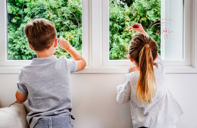 With Kitpas Crayons - 6 Colors, two children create vibrant drawings on window panes, showcasing the lush green foliage beyond. One child, dressed in a gray shirt with short hair, and the other wearing a striped top with long hair tied back, both concentrate intently on their non-toxic artwork.