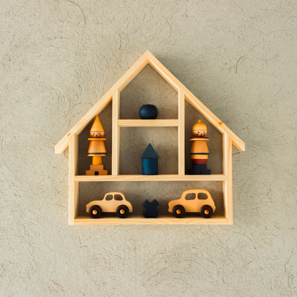 A wooden house-shaped wall shelf displays various wooden items, including two toy cars, a blue tree, a blue ball, a blue cube, and two cone-shaped figures resembling people, all arranged neatly within the solid wood shelves. This delightful piece is perfect storage for children's toys and trinkets: Wooden Shelf No. 21.