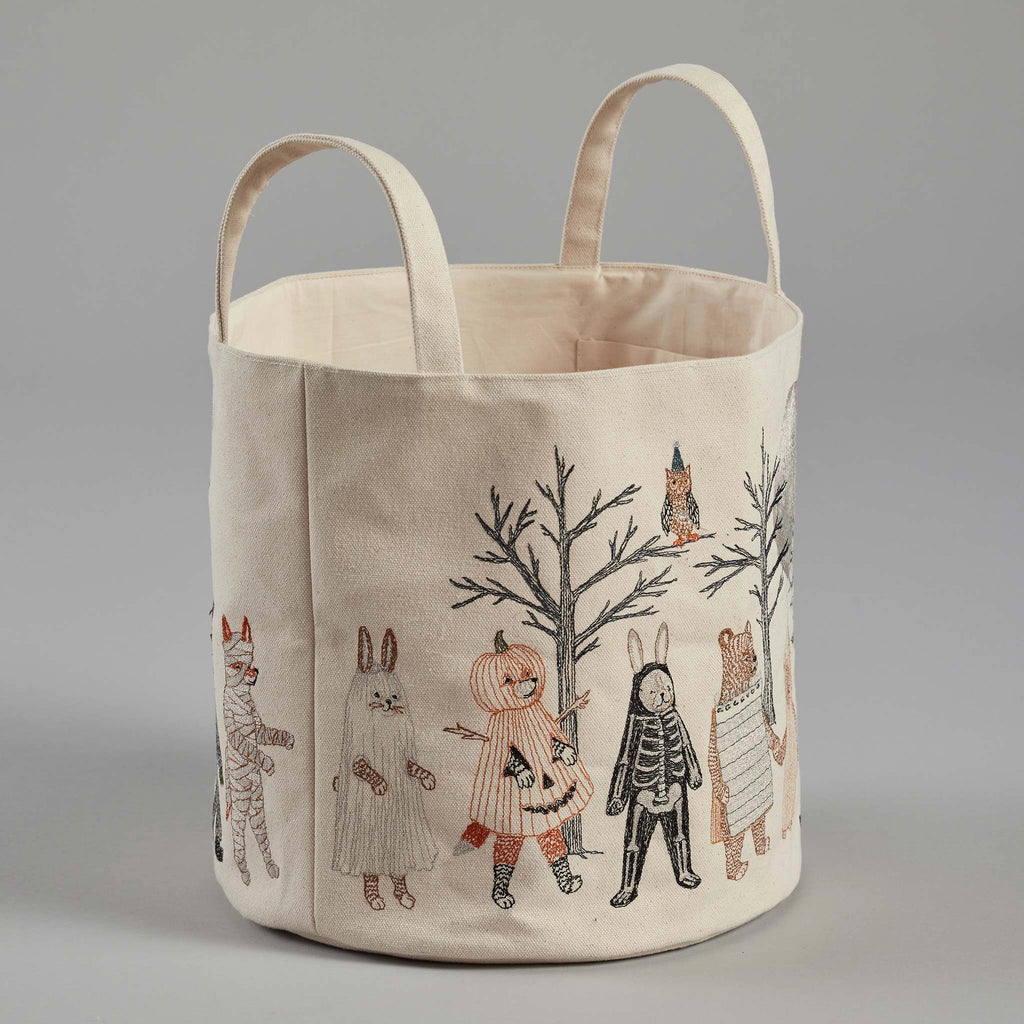 The Coral & Tusk Full Moon Masquerade Bucket is a fabric storage basket with two handles, featuring illustrated designs of children in various Halloween costumes—including a mummy, ghost, pumpkin, skeleton, and animal outfits—set against a light gray background adorned with bare trees and an owl.