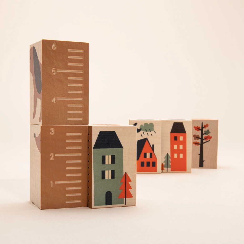 A series of Uncle Goose Environments Neighborhood Blocks arranged to depict a quaint village scene with houses, trees, and a ruler design on one block, against a plain light background.