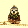 The Happy Sloth Necklace features a handcrafted porcelain pendant shaped like a smiling sloth with green leaf accents around its neck. The pendant has a golden loop at the top and is attached to a delicate 24k gold plated steel chain, lying against a pale yellow background.