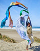 A joyful child leaps on a sandy beach, waving Sarah's Silk Giant Ocean Playsilk in the air. The scene, with the ocean and clear blue sky as a backdrop, is vibrant and lively, inviting imaginative play that knows no limits.