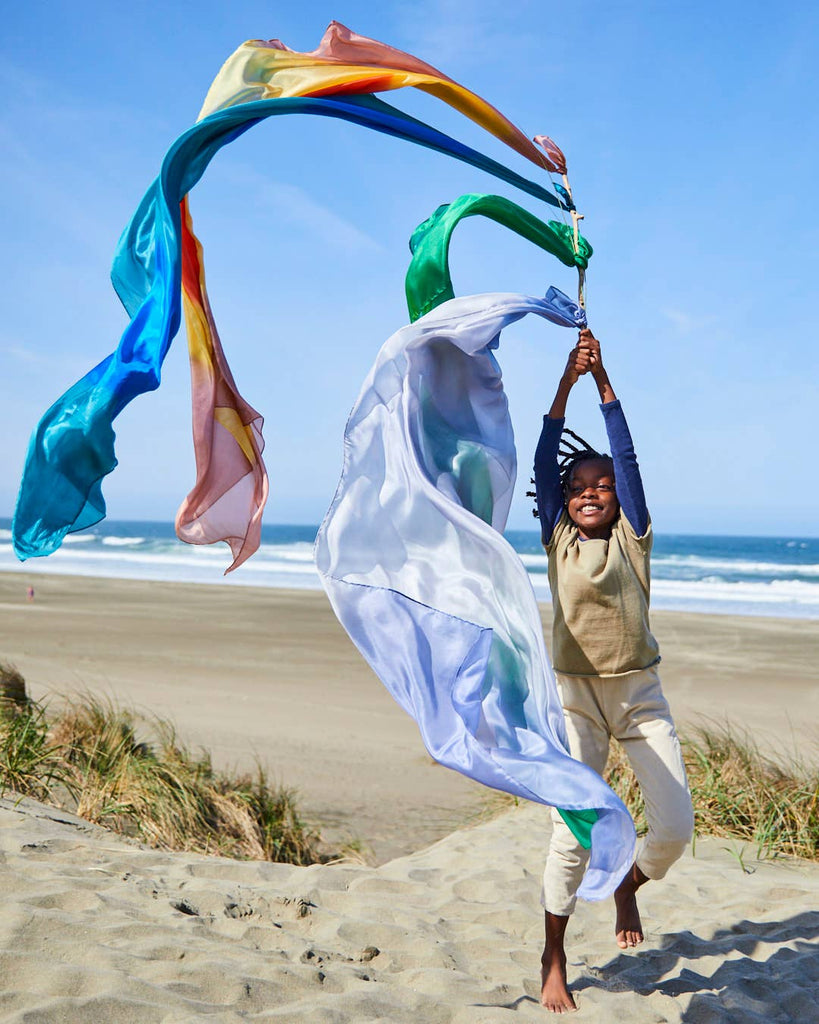 On a sandy beach under a clear blue sky, a joyful child leaps and twirls with Sarah's Silk Giant Forest Playsilk. The vibrant fabric flows gracefully in the air as ocean waves roll in the background, enhancing imaginative play and creating a lively and cheerful scene.