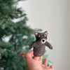 In a hand, a small wool finger puppet shaped like a raccoon is held. Made of dark felt, it stands out as a charming handmade piece. In the background, a blurred Christmas tree with green branches can be seen.