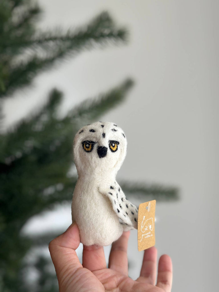 A hand showcases the Snowy Owl Finger Puppet, a small white felt owl with black spots and striking yellow eyes from the 2024 Finger Puppet Collection. It has a tag attached to its wing, while in the background, a green Christmas tree adds charm to this handmade piece.