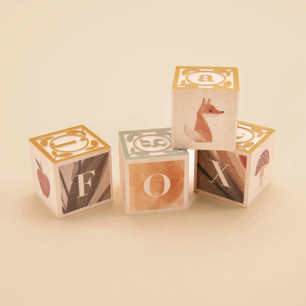 Four Uncle Goose Watercolor ABC Blocks are creatively stacked, spelling "FOX" with artistic letters. Featuring images of a fox, an apple, and a mushroom, these educational toys are crafted from sustainable basswood in earthy tones against a minimalist neutral background.