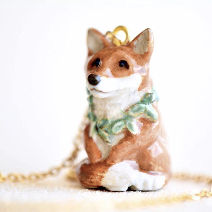 A delicate Fox Necklace shaped like a brown and white corgi dog, adorned with a green wreath around its neck, is displayed against a plain background. The hand-painted pendant is attached to a gold plated chain.