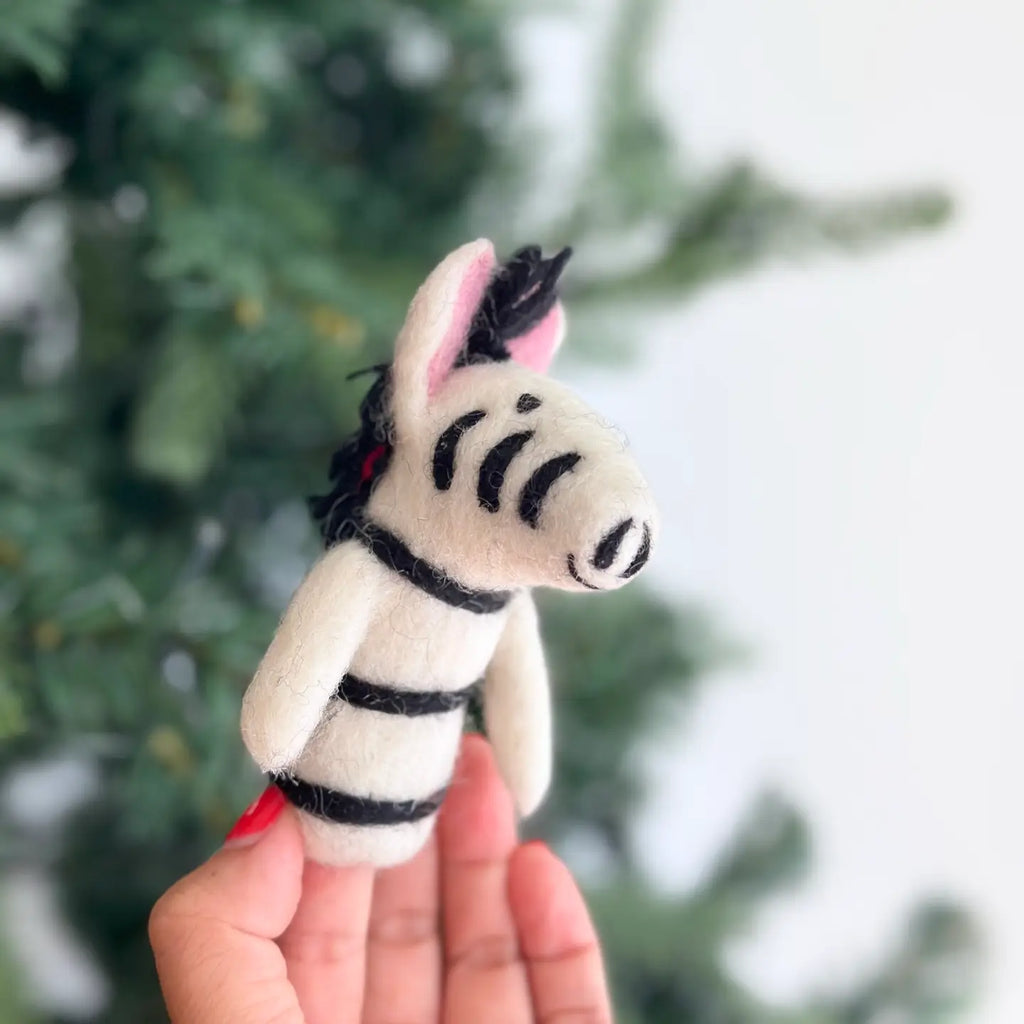 A hand with red-painted nails holds the Zebra Finger Puppet, a small, needle-felted toy featuring black stripes, a white body, and pink inner ears, against a blurred green background that looks like trees—ideal for wild animal enthusiasts and a charming addition to any storytelling toy collection.