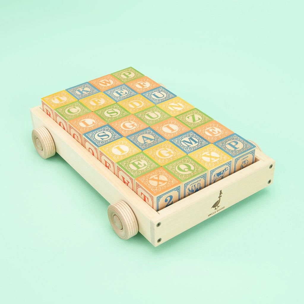 A handmade Uncle Goose Classic ABC Blocks with Wagon carries neatly arranged embossed ABC blocks in vibrant colors, featuring letters, numbers, and animal images on a light turquoise background.