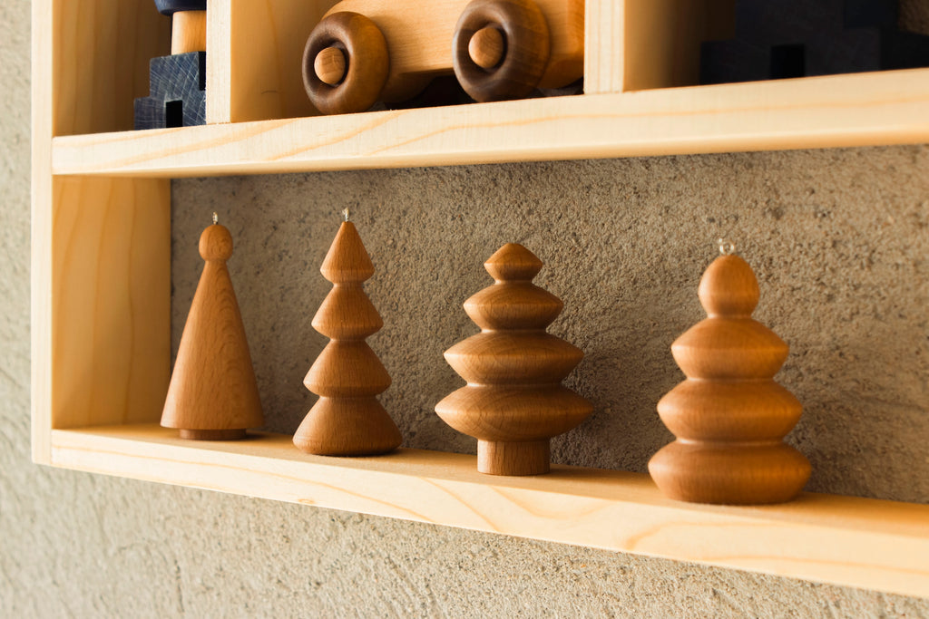 A Wooden Shelf No. 21 displays four handcrafted wooden Christmas tree decorations and a toy train. Each tree boasts a unique design, from simple geometric shapes to intricate layered forms. Set against a textured wall, this charming scene makes for perfect storage shelves in children's rooms.