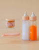 The Bottled Milk and Juice Set by Tiny Harlow includes two magic bottles, one filled with white liquid and the other with orange. Next to them is a small jar labeled "Tiny Tummies Peach Jelly," ideal for feeding dolls. A pink spoon lies nearby on a clear, flat surface.