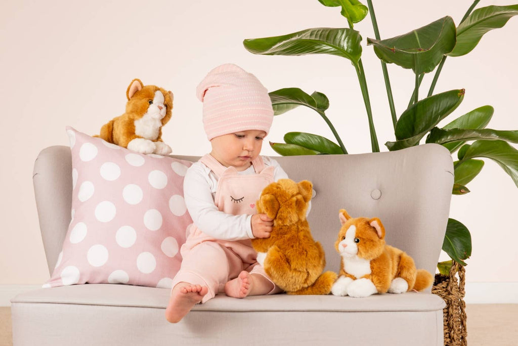 A baby wearing a pink beanie and overalls sits on a gray couch adorned with white polka dot pillows, surrounded by high-quality plush toys from the Hermann Teddy Collection. The baby is holding the Teddy Hermann Orange Laying Cat Stuffed Animal, with green plants in the background.