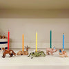 A whimsical display featuring the Cricket Cake Topper, a hand-painted porcelain animal figurine (a fox, two deer, and a frog) each wearing a small party hat and balancing colorful birthday candles on their backs, arranged on