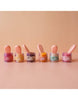 A row of six cheerful jars labeled "Tiny Harlow" stands ready, each containing a magical spoon. From left to right, the jars overflow with vibrant colors: pink, yellow, orange, green, purple, and another pink. The gentle peach background embraces this charming scene made for Tiny Tummies - Banana Jelly Food.