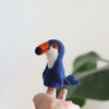 A hand holding the Toucan Finger Puppet, a small, needle-felted creation of a blue bird with an orange beak. Handmade from 100% wool, this delightful finger puppet stands out against the blurred background of green foliage.