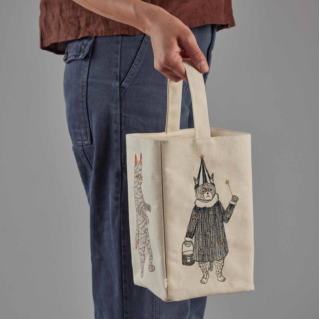 A person is holding the Coral & Tusk Halloween Costumes Bag, a cream-colored fabric bag adorned with embroidered cat designs ideal for trick-or-treating. One cat is dressed as a wizard with a pointy hat and wand, while another in the background appears to be wrapped like a mummy. The person is wearing blue jeans and a brown top, showcasing these adorable costumed critters.