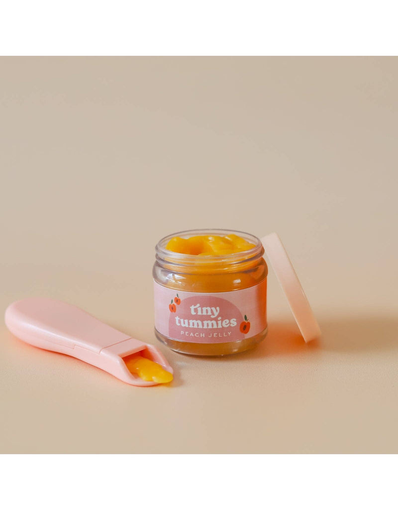 A small open jar of "Tiny Tummies - Peach jelly food" by Tiny Harlow, topped with a peach-colored lid, rests on a light surface. Beside it lies a magic spoon, holding just the right amount of jelly for doll food adventures.