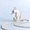 A small, gold Arctic Seahorse Necklace with a textured surface, hanging from a delicate gold plated chain, set against a soft blue background with a light dusting of snow-like particles.