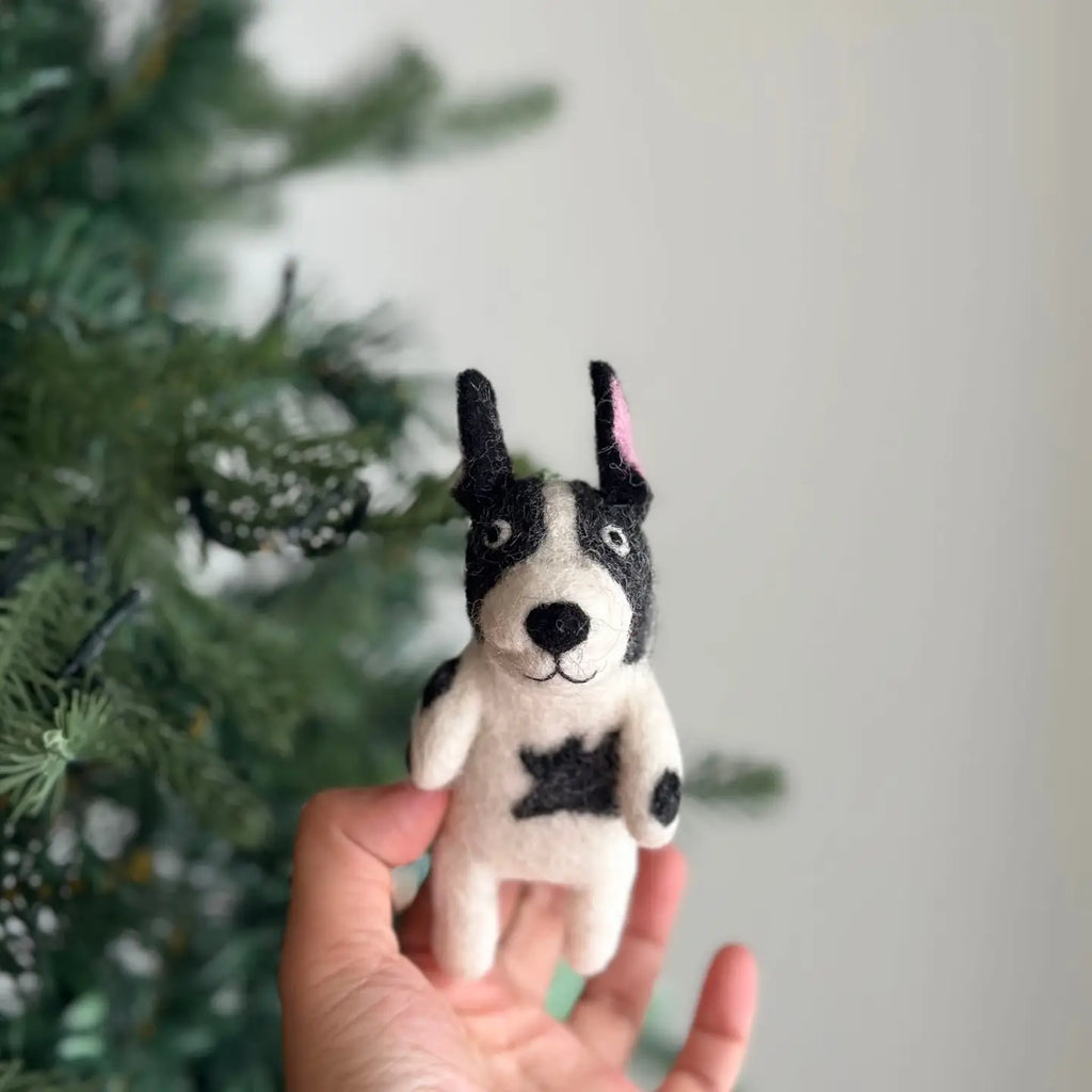 A hand is holding the adorable French Bull Dog Finger Puppet, a black and white handcrafted figurine made of 100% wool. The background showcases a blurred green Christmas tree adorned with decorations. Handmade in Nepal, this charming dog has black ears, one of which is slightly flopped.