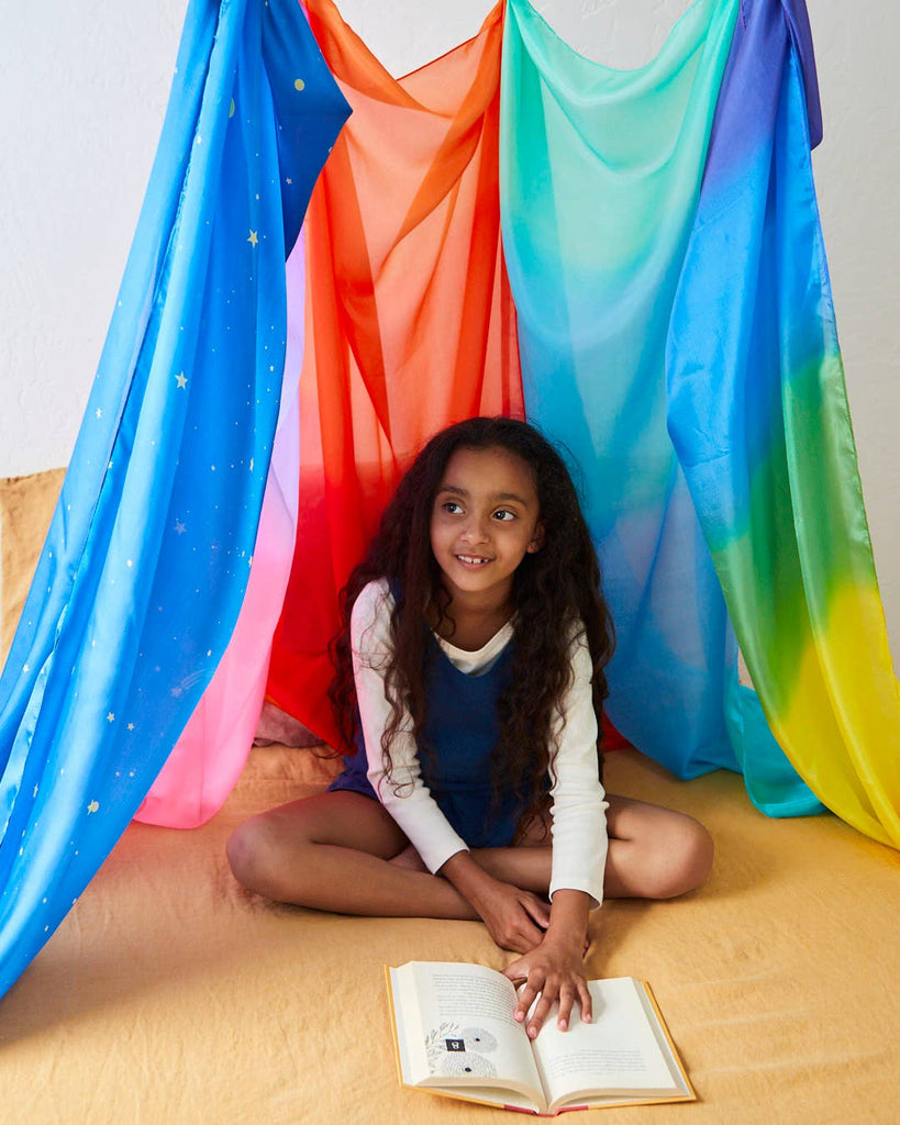 A young girl with long curly hair sits cross-legged, smiling inside a colorful tent made of draped, star-patterned Sarah's Silk Giant Forest Playsilks. She wears a white shirt and blue dress and has an open book on her lap. The setting exudes a cozy vibe perfect for imaginative play.