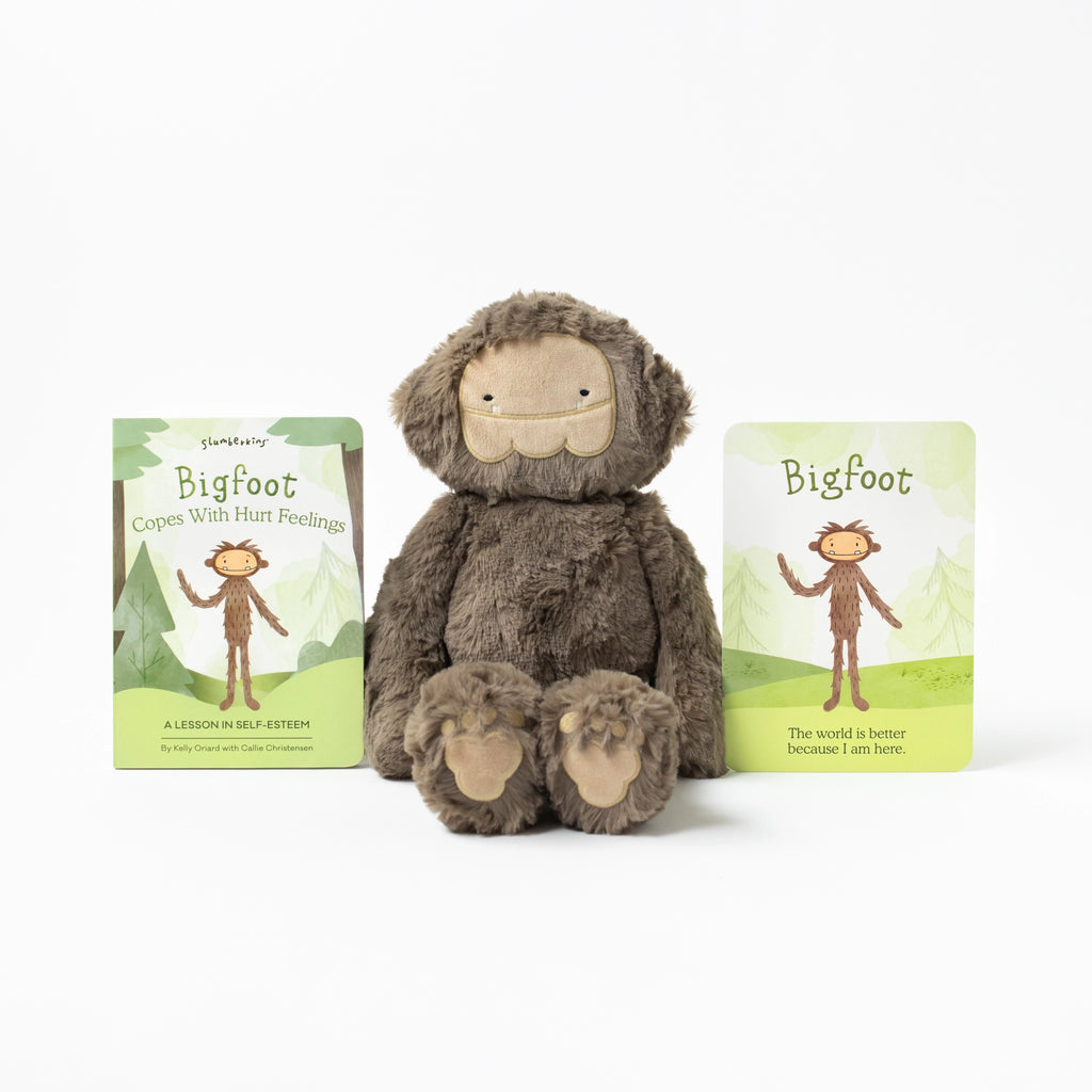 A Slumberkins Bigfoot Kin + Lesson Book On Self Esteem stuffed with hypoallergenic fiberfill sits between two children's books titled "Bigfoot Copes with Hurt Feelings," featuring a happy, friendly design on a white background.