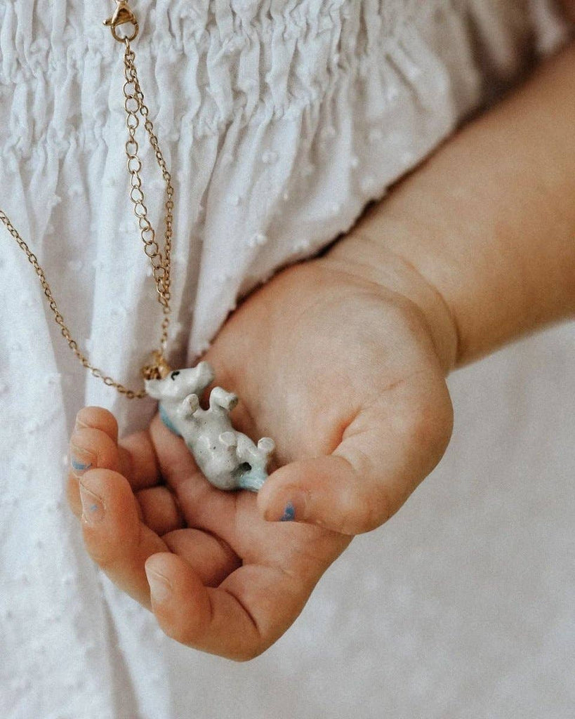 A child's hand gently holds a whimsical wearable—a Galaxy Unicorn Necklace with a small silver hippopotamus pendant—against a white, textured fabric background.