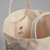 Close-up of the interior of the Coral & Tusk Full Moon Masquerade Bucket, a beige tote bag resembling a canvas container. The image reveals a small inner pocket adorned with a white label featuring an embroidered red coral design, a small black ship, and the text "CORAL & TUSK." The background is plain and grey.