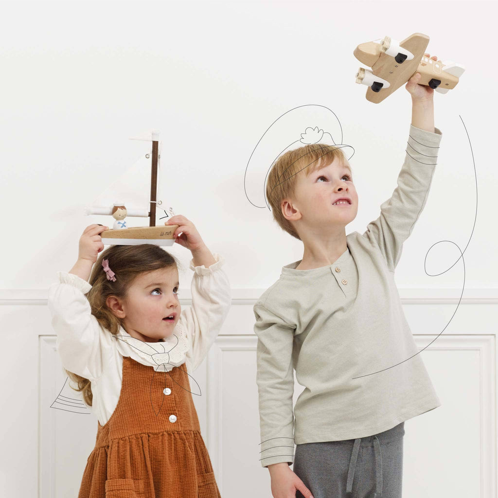 Two children play indoors with handcrafted toys; a girl in a brown dress holds a Wooden Sailing Boat & Captain over her head, while a boy in a grey top lifts a toy airplane. Both are smiling and looking upward, surrounded by whimsical line illustrations that spark their cognitive skills.