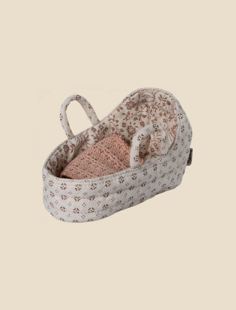 A small, oval-shaped Maileg Carry Cot, Baby with handles, featuring a floral pattern on the exterior and interior lining. Inside the basket is a knitted pink blanket, neatly folded. This versatile carrycot can also double as a portable bunny bed. The background is a plain, light beige color.