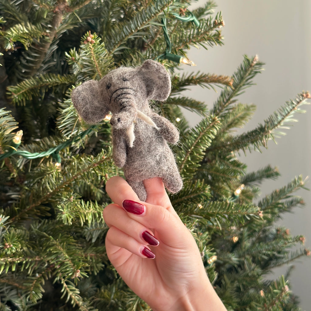 In front of a decorated Christmas tree adorned with twinkling white lights, a hand with red-painted nails holds a small, gray Elephant Finger Puppet.