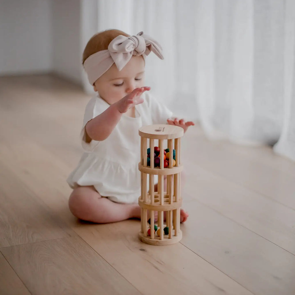 A baby wearing a white outfit and a large bow headband sits on a wooden floor, playing with the Wooden Rain Maker made from non-toxic materials. The toy contains colorful balls inside, and the baby is holding it with one hand while reaching for it with the other. White curtains are in the background.