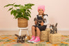 A young girl in a pink beanie and pink sneakers sits on a wicker basket, holding a high-quality Teddy Hermann Standing Tabby Cat stuffed animal. She is surrounded by two other cuddly cats from the Hermann Teddy Collection. A potted plant is on a stool to her left, all set against a light-colored wall and a patterned rug.