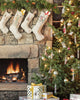 A cozy living room radiates holiday cheer, featuring a stone fireplace with four stockings hanging above. Beside it stands a Christmas tree, proudly adorned with lights and ornaments including the charming Bear with Tinsel Ornament. Wrapped presents in festive paper are placed near the tree, completing the joyful scene.