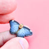 Close-up of a hand holding a small, detailed model of a blue and black butterfly with white spots along the edges of its wings, set against a pink background. The Blue Butterfly Necklace is primarily blue with black bordering and a beige body.