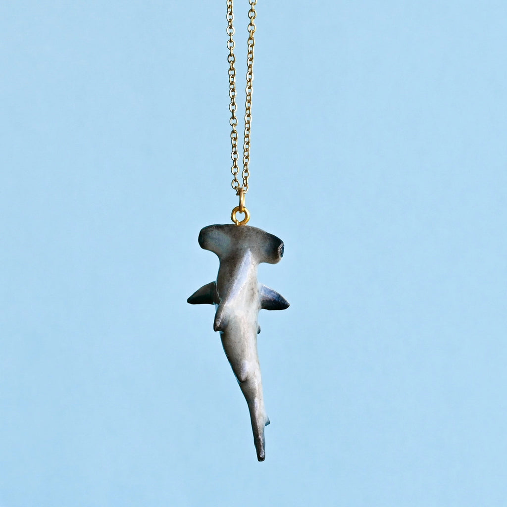 A Hammerhead Shark Necklace hangs on a delicate gold chain against a light blue background. The whimsical wearable, crafted from high quality porcelain, captures the unique shape of the shark with a broad, flattened head and streamlined body.