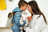 A young boy with a backpack and a Slumberkins Otter Kin + Lesson Book - Family Bonding touching noses affectionately with his smiling mother in a warmly lit room, both wearing jackets.