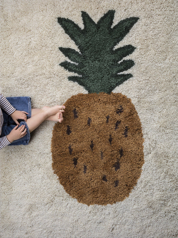 A child sits next to a large Ferm Living Fruiticana Tufted Pineapple Rug on a light carpeted floor, with only the lower part of their body visible. The rug features a textured brown base with black details and a green.