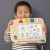 A child wearing a striped shirt holds a Wooden Elephant Magic Drawing Board with magnetic stamps. The board features colorful buttons and displays doodles of a sun, trees, numbers, and a house on its screen against a plain gray wall in the background.