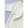 Close-up of a cream-colored organic cotton Baby Jumpsuit with short sleeves, snapped open to show a detail of the side snaps. The garment is laid out on a blue surface.