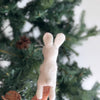 A hand is holding a small Bunny Finger Puppet, handmade in Nepal, in front of a decorated Christmas tree. The puppet is facing away from the camera, showing its back and pointed ears. The tree is adorned with pinecones and green foliage.