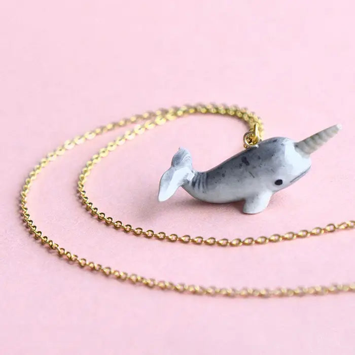 A delicate gold necklace featuring a hand-painted narwhal charm set against a soft pink background. This whimsical narwhal charm is gray with a white tusk, black eyes, and a slight smile, making this Narwhal Necklace an enchanting piece.