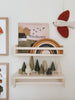 A minimalist children's room features wooden shelves displaying imaginative play toys. The upper shelf holds a book titled "Birdsong" and a wooden rainbow, while the lower shelf showcases SABO concept toys and the Wooden Forest Set. A fabric bird on the wall enhances the earthy, calming atmosphere.