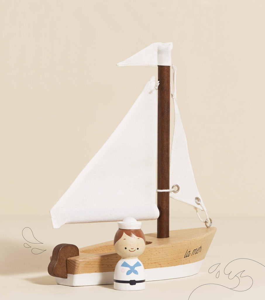 The Wooden Sailing Boat & Captain features a meticulously crafted toy sailboat with white sails and a small sailor figure at the helm. The phrase "la mer" embellishes its side, beckoning children to engage in imaginative play. Positioned against a light beige backdrop accentuated with simple wave illustrations, it perfectly embodies the adventurous spirit of the open sea.