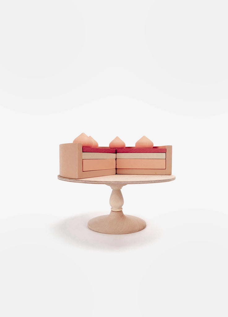 A Handmade Strawberry Layer Cake On A Stand with stylized pink fondant, displayed on a wooden stand against a soft white background. The cake features three visible tiers and decorative top elements, coated in non-toxic paint.