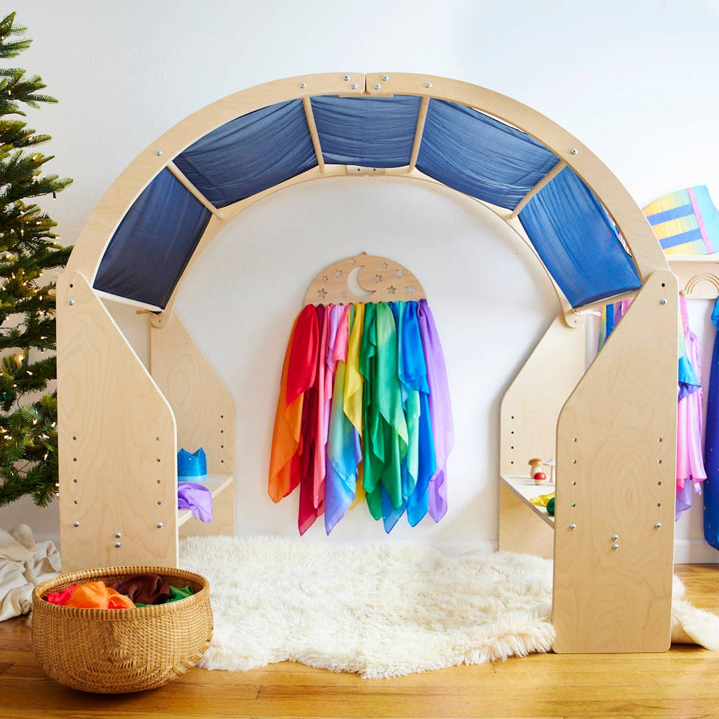 A wooden play arch adorned with vibrant fabric drapes is surrounded by a plush white rug, creating an inviting space for imaginative play. Sarah's Silk Giant Midnight Playsilk hangs in the center, adding a touch of magic, while a basket of toys rests nearby. A partially visible Christmas tree on the left enhances the setting for endless open-ended play adventures.