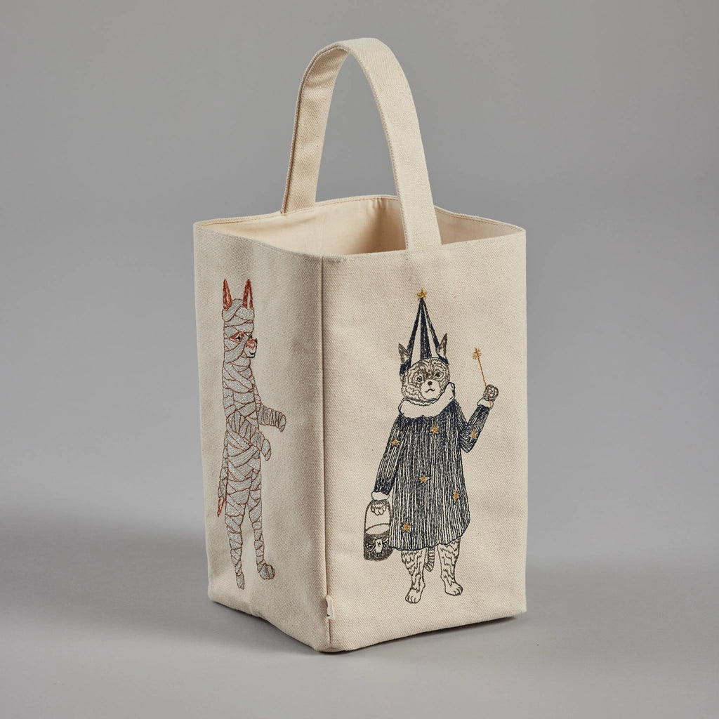 A beige fabric tote bag with a handle stands upright against a gray background. The Coral & Tusk Halloween Costumes Bag features an embroidered design of costumed critters: two cats dressed in festive outfits, one wrapped as a mummy and the other in a wizard outfit holding a wand and lantern.