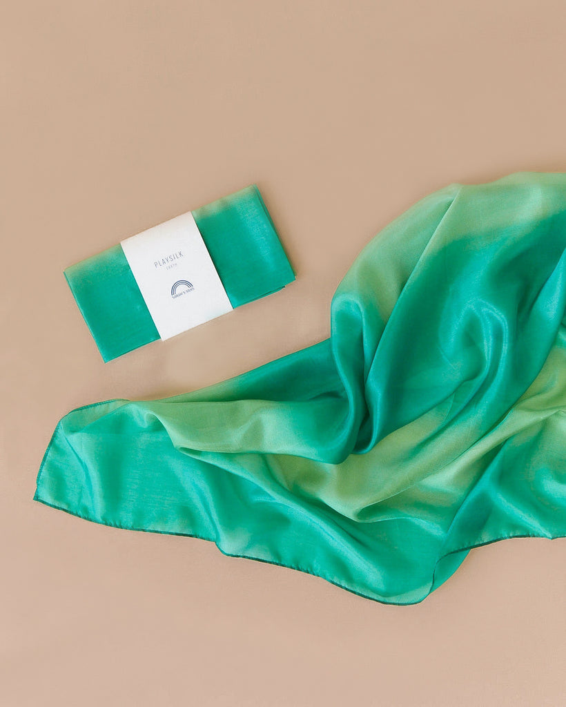 Sarah's Silk Enchanted Playsilk in the Forest design, featuring shades of green and turquoise, is elegantly spread on a beige surface. To the left, a similar playsilk for imaginative play is folded and wrapped in white packaging.